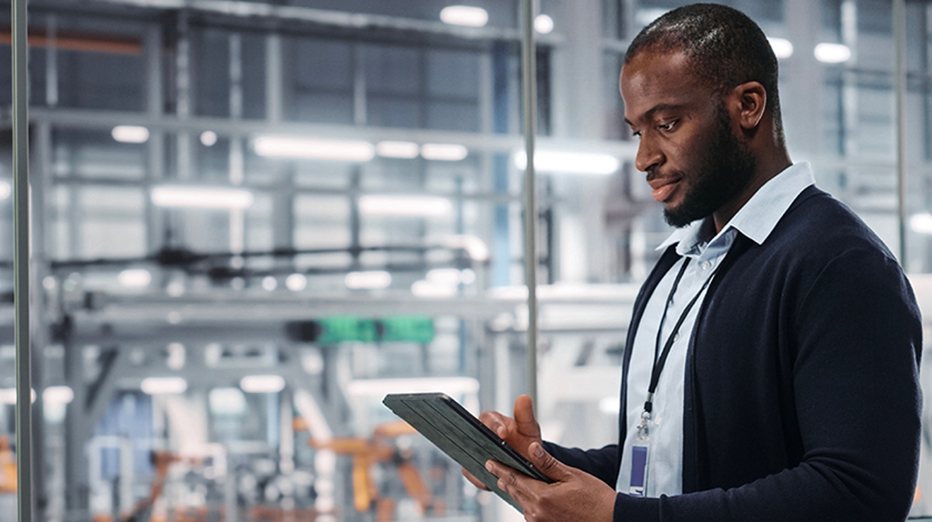 A young professional works on a tablet. A production facility is visible through the window behind him.