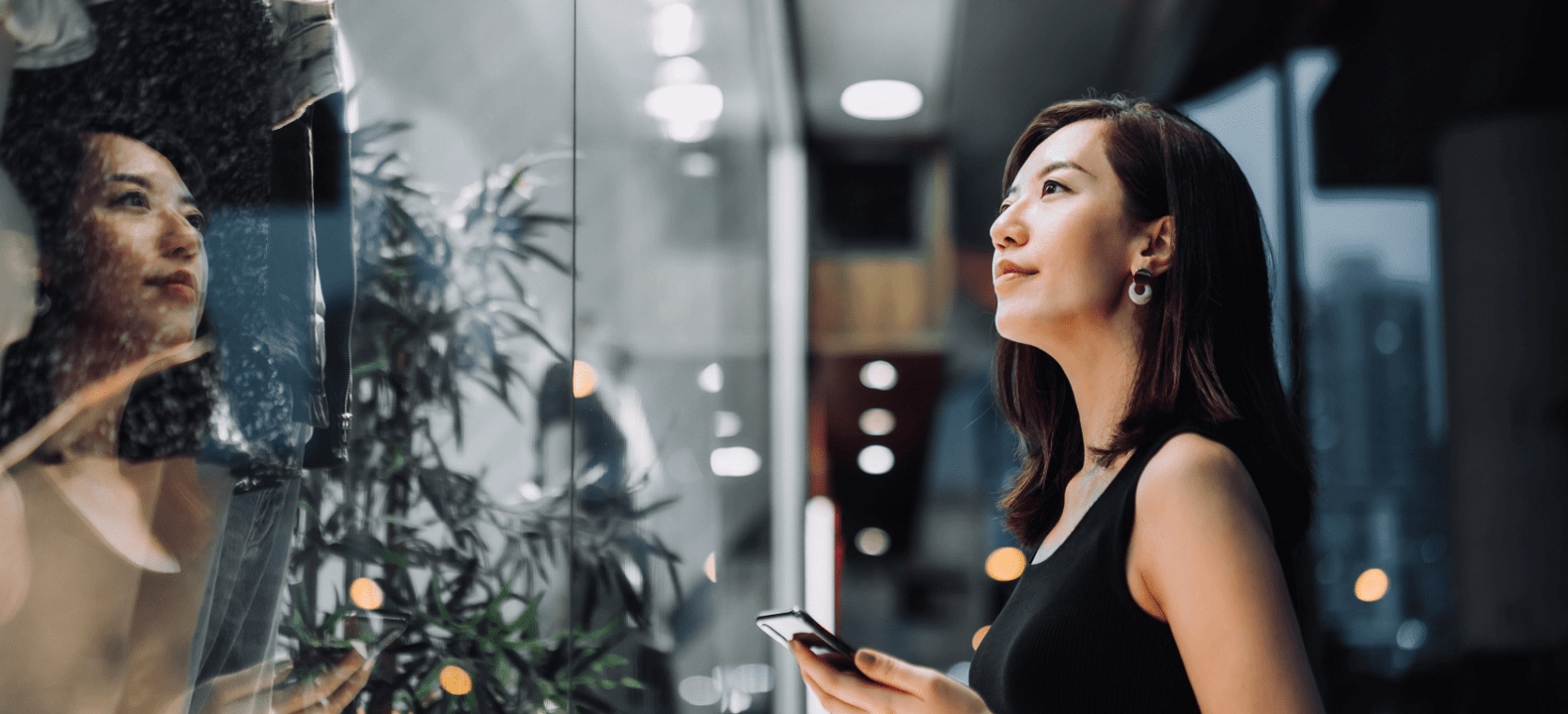 A woman gazes into a store window, smartphone in hand.