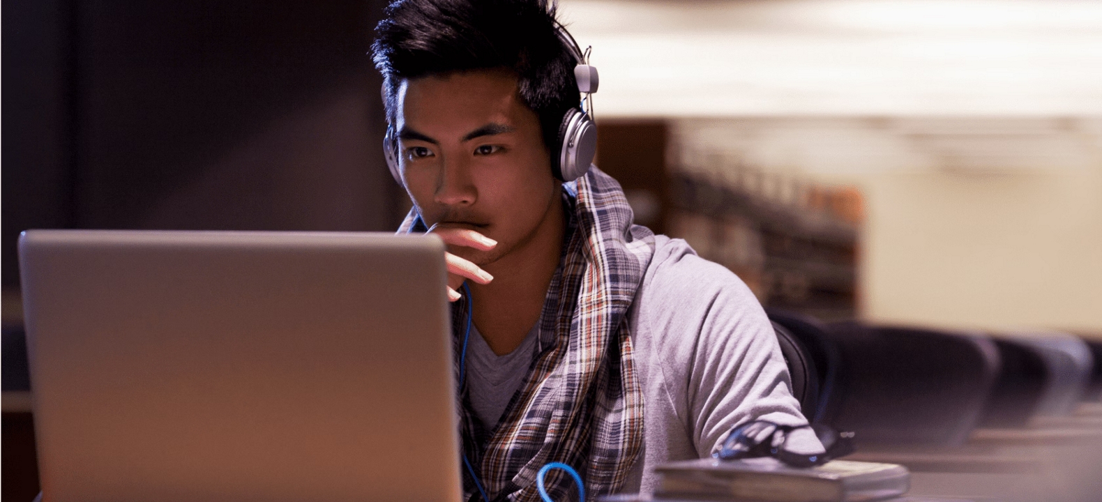 A college student wearing headphones studies on his laptop while seated in a library.
