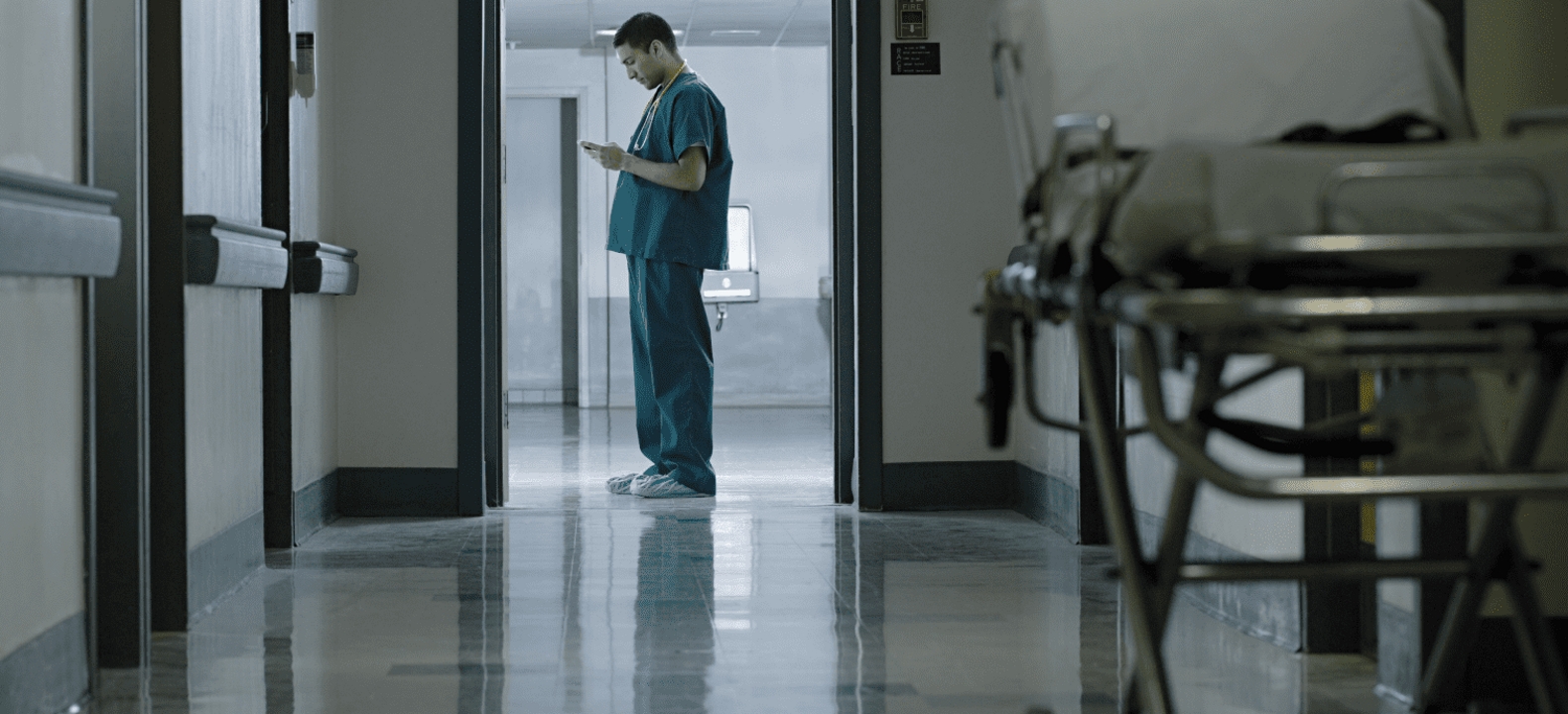 A healthcare professional on his smartphone at the end of a hospital hallway.
