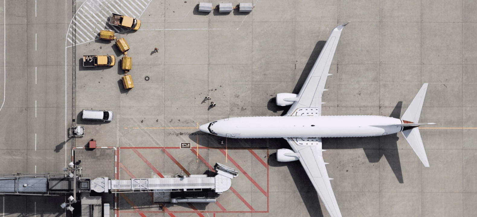 An aerial view of an airport gate, including a jet bridge, trucks, and airliner.