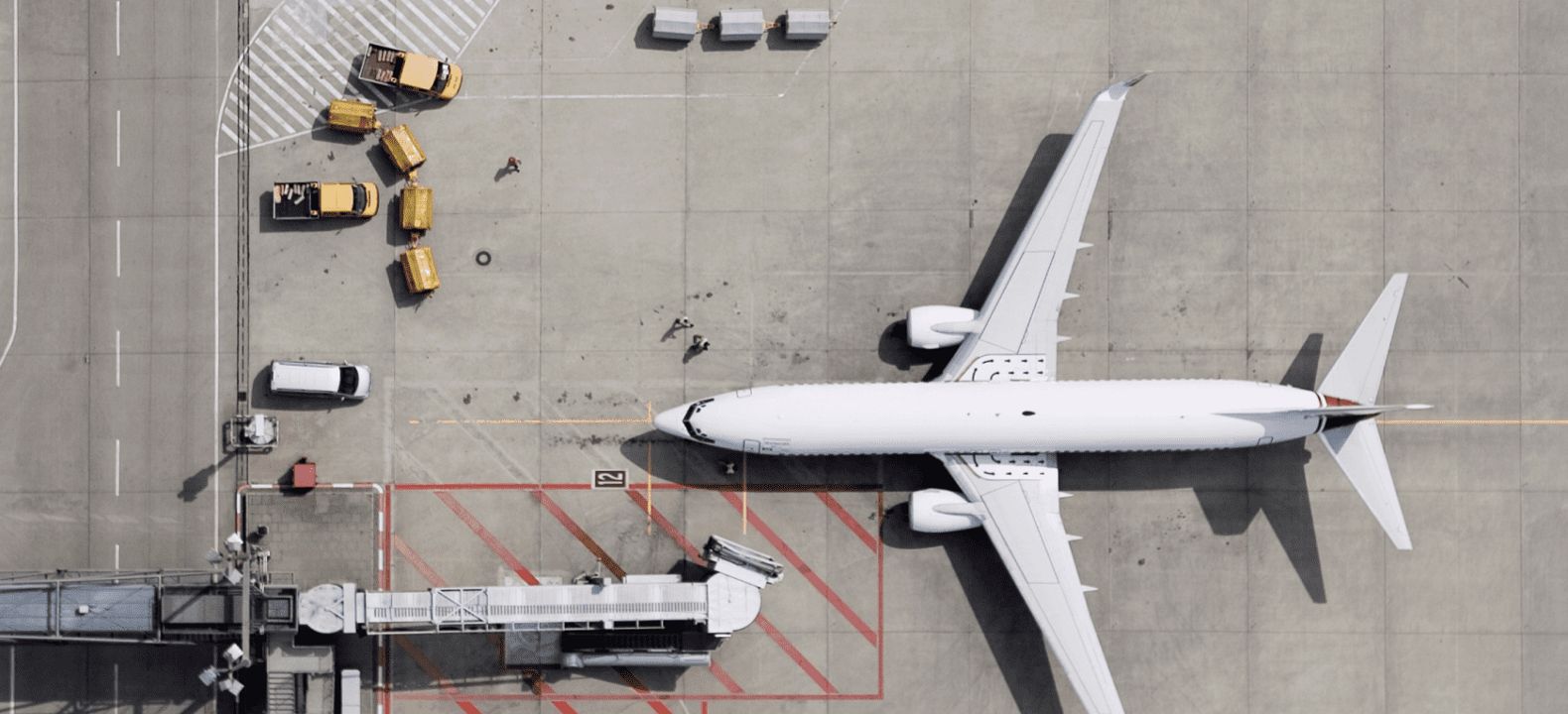 An aerial view of an airport gate, including a jet bridge, trucks, and airliner.