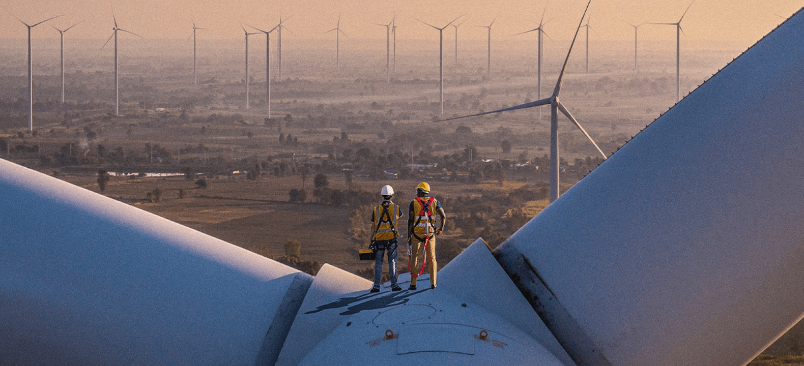 Two construction workers stand atop of a windmill looking out on a horizon filled with other windmills.