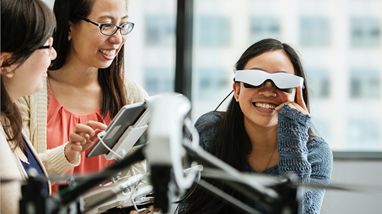 Two female students look at a third smiling student who holds a VR viewer to her head.