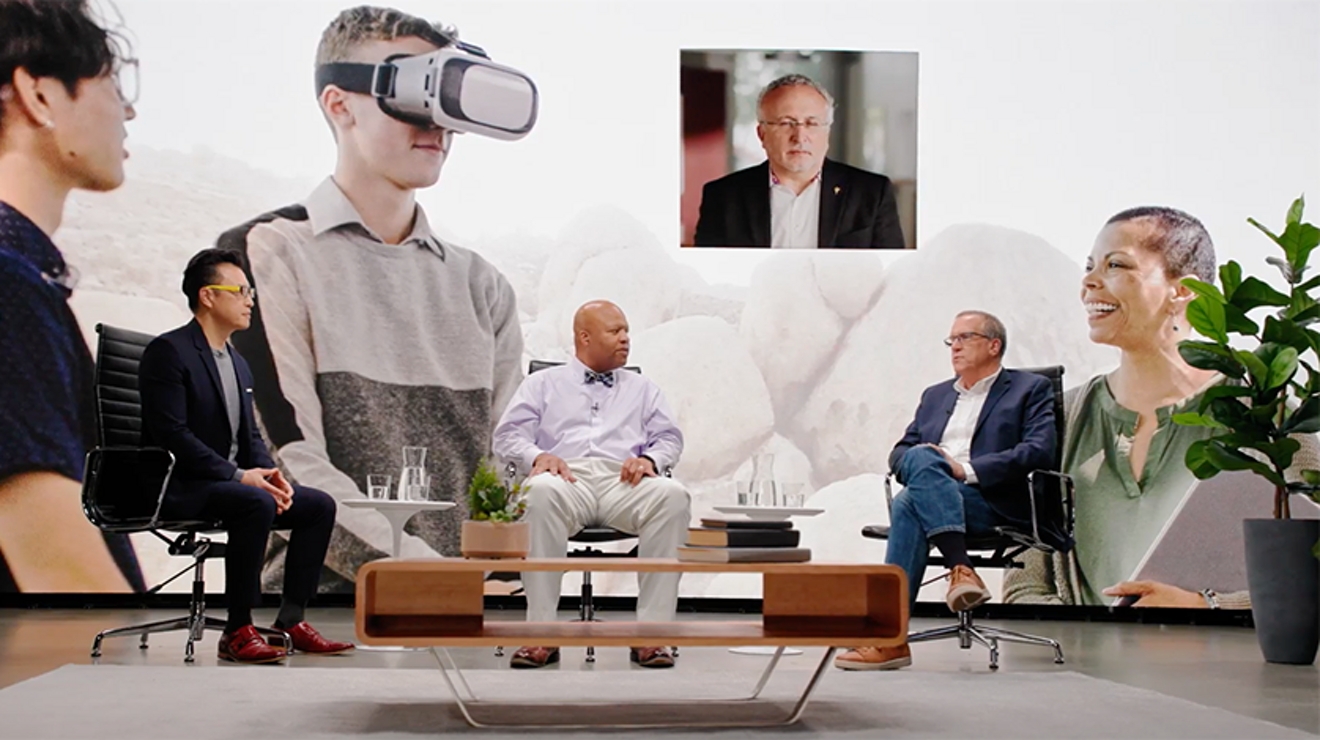 Two men sit in chairs on stage with an oversized image of a student in VR gear behind them Watch the full workshop video.