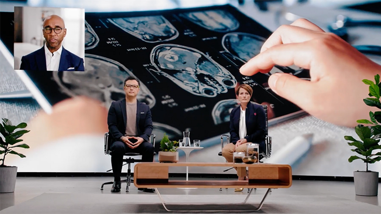 Two people sit in chairs on stage with an oversized image of a tablet showing MRI results.