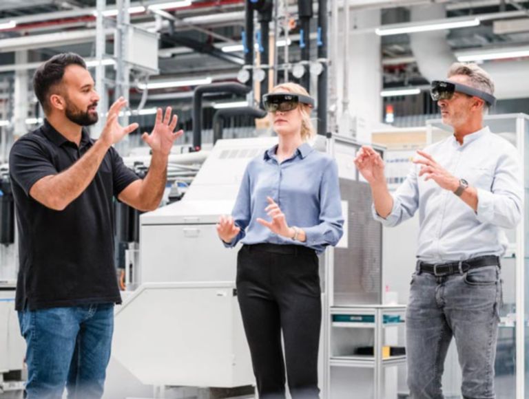 Person in an industrial setting instructs two other people while they’re wearing VR glasses