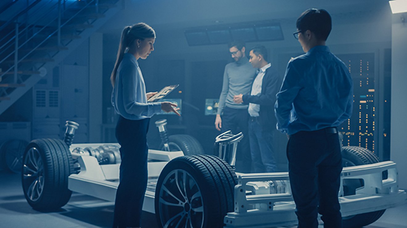 Four professionals, one with a tablet in hand, examine a vehicle frame.