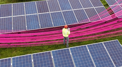 Person in a hardhat and safety vest inspects solar panels while holding a clipboard.