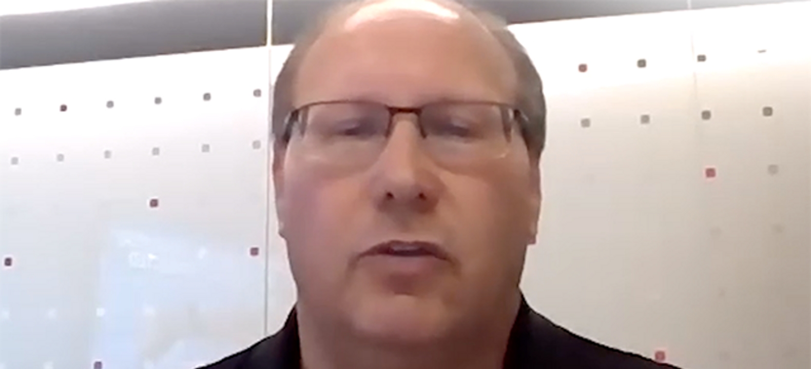 Closeup of bald man with glasses and dark shirt sits in front of a white background