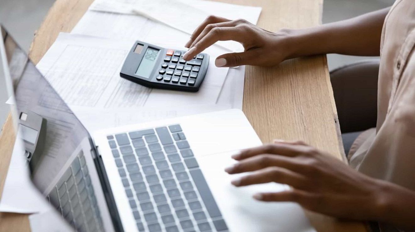 A business professional works on a laptop with one hand and on a calculator with the other.