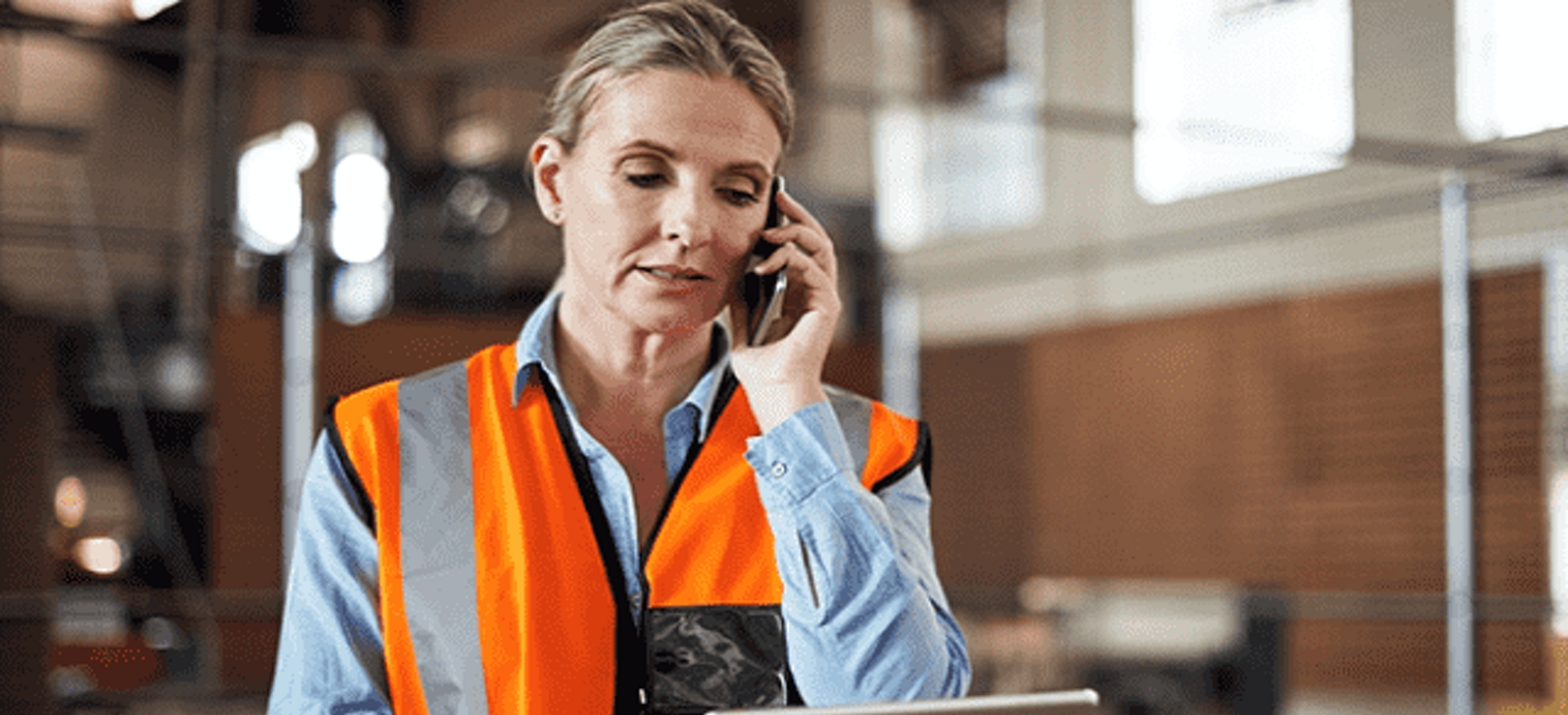 Worker in safety vest leans on warehouse boxes while looking at tablet and talking on smartphone.
