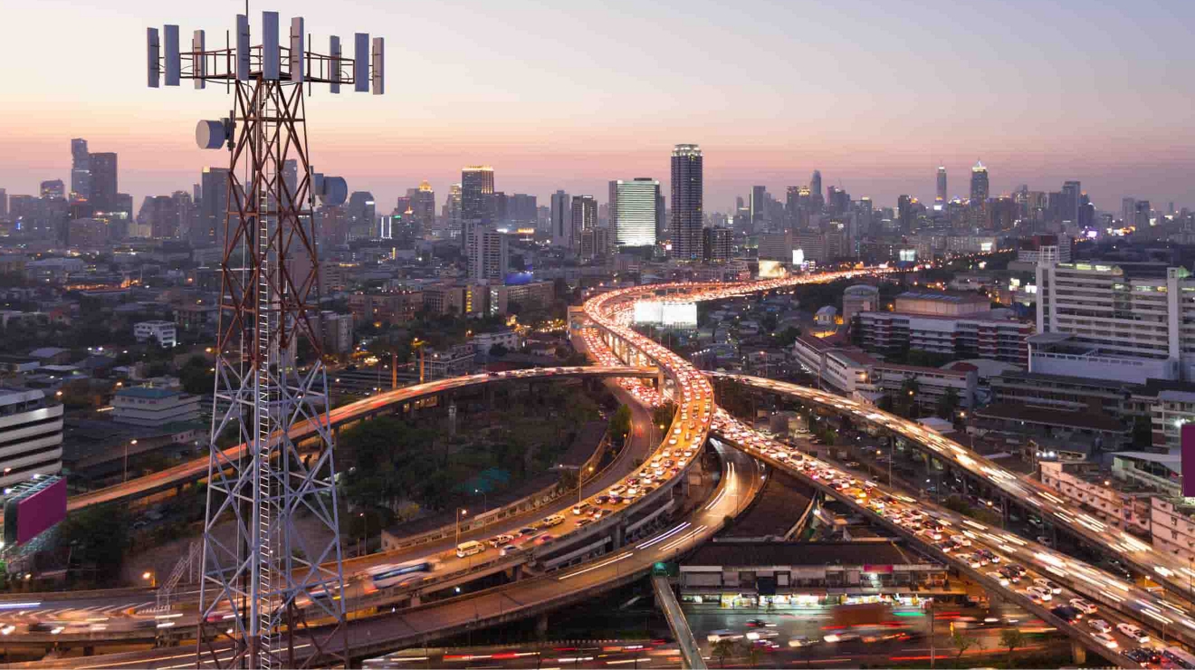 An aerial view of busy freeways with a cell tower in the foreground and a city in the distance.