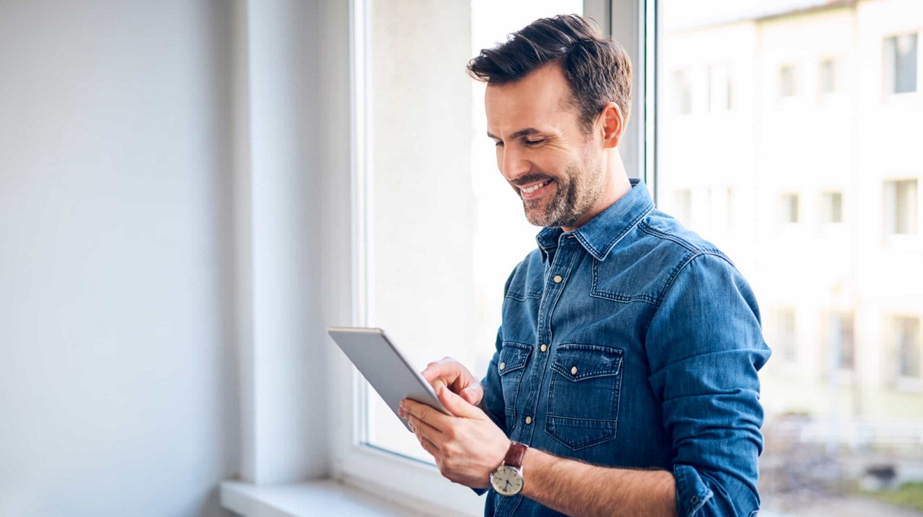 A smiling business professional leans against an office windowsill and types on a connected tablet.