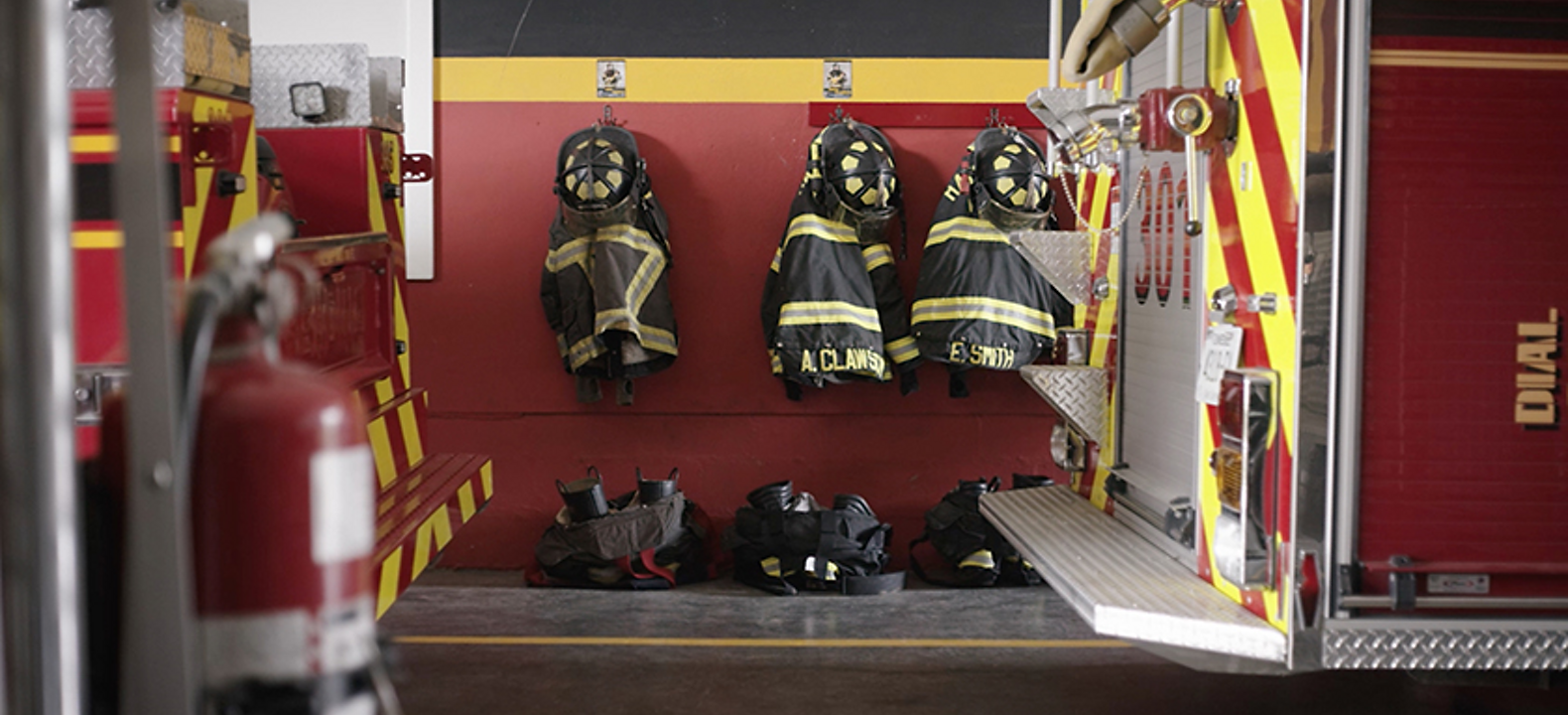 The back of a fire truck inside a fire station with firefighter gear hanging on a wall and boots on the floor
