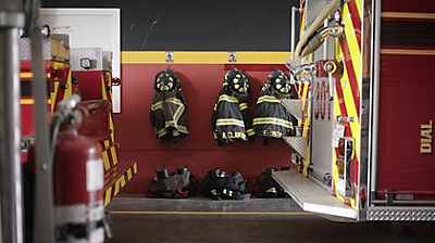 The back of a fire truck inside a fire station with firefighter gear hanging on a wall and boots on the floor