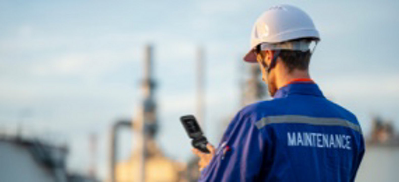 Man in white hard hat with blue maintenance uniform looks down at smartphone