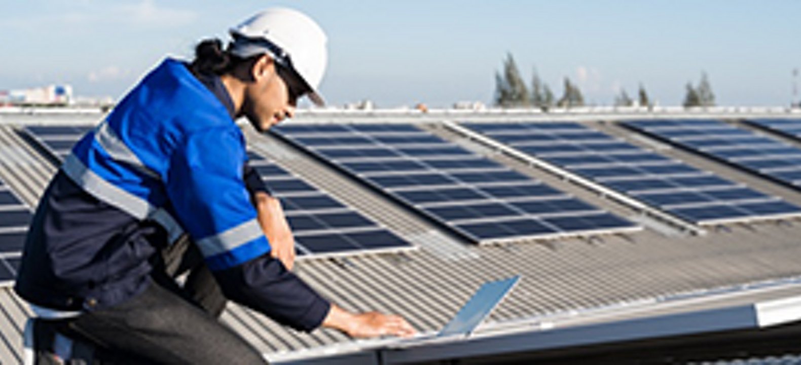 Man in blue vest with white hard hat works on device outside next to solar panels