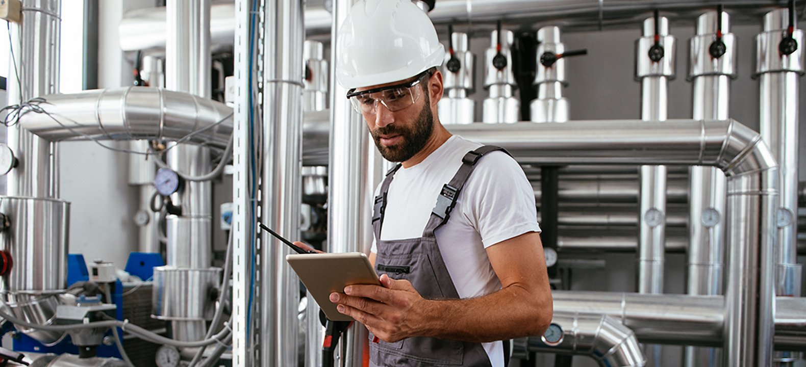 Man in overalls with white hard hat stand in front of pipes looking down at a tablet in his hands