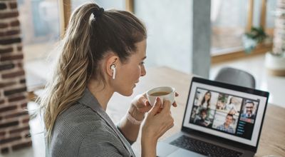 Woman with ponytail and ear buds sits looking at video conference on screen