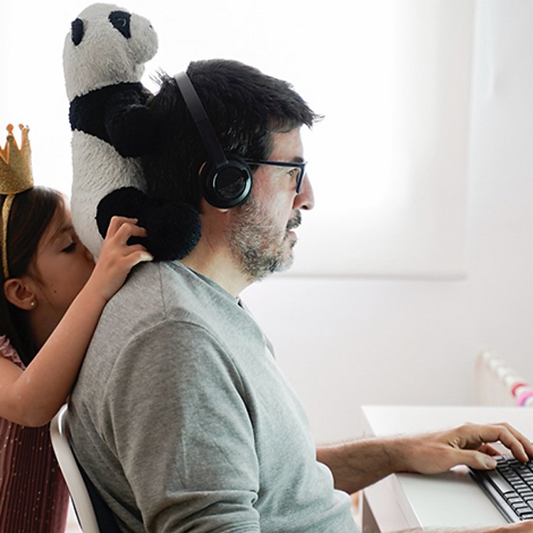 Man sitting at computer with headphones on while child holds panda bear on his neck behind him.