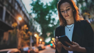 A financial services professional checks her smartphone while walking on a city street.
