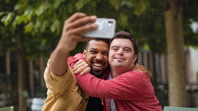 Two friends laugh as they take a selfie together in a sunlit garden.
