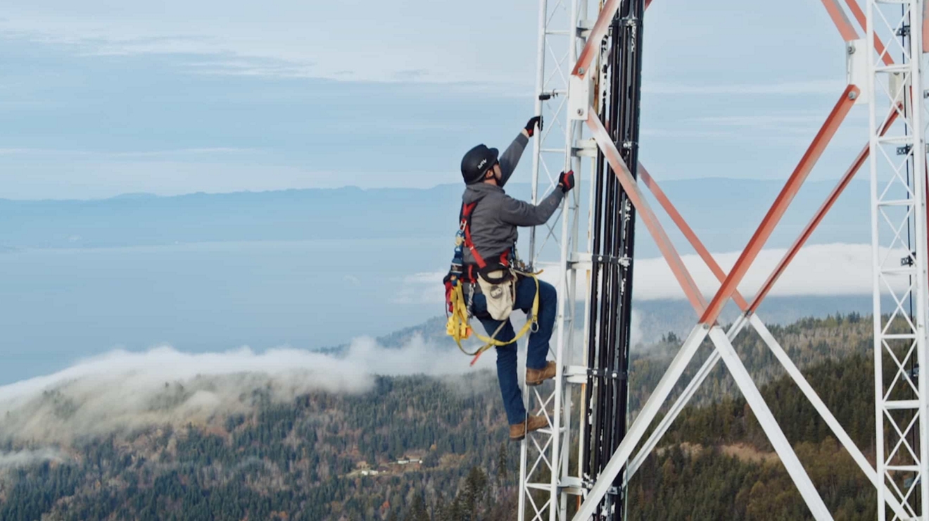A tower worker wearing safety gear climbs a cell tower in a rural location.