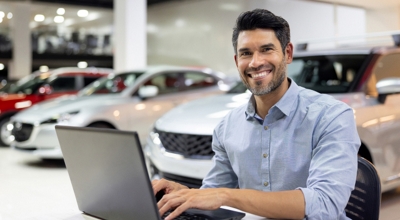 A smiling salesperson at an auto dealership provides efficient customer service via their laptop.