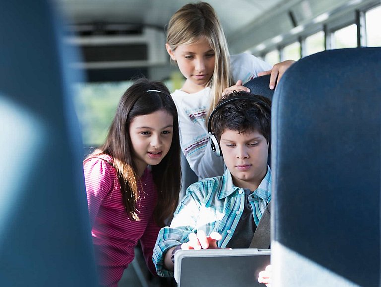 Three grade-school students ride a Wi-Fi-connected school bus and share their work on a mobile device. 