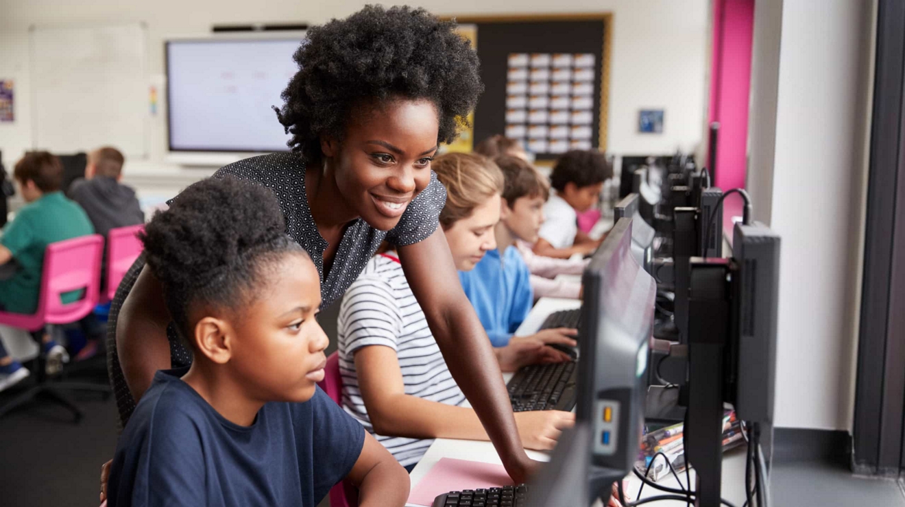 Students work independently on desktop computers while a teacher checks their work.