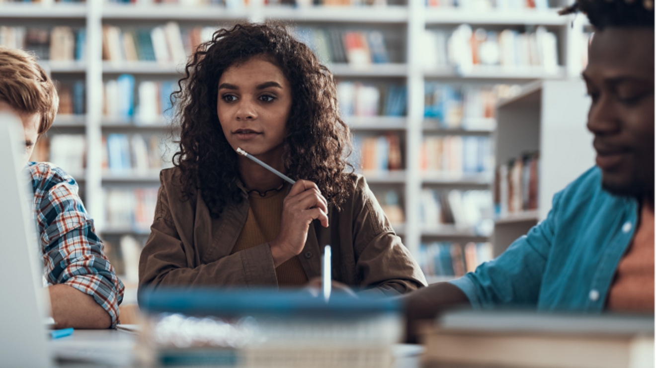 Students work together in a library with a mix of pencils, paper, and a laptop computer.