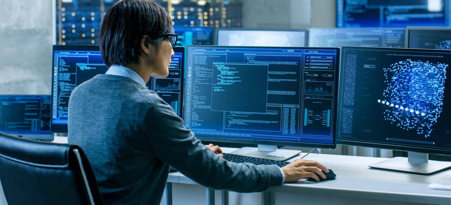 An IT professional in a server room at work in front of a bank of monitors.