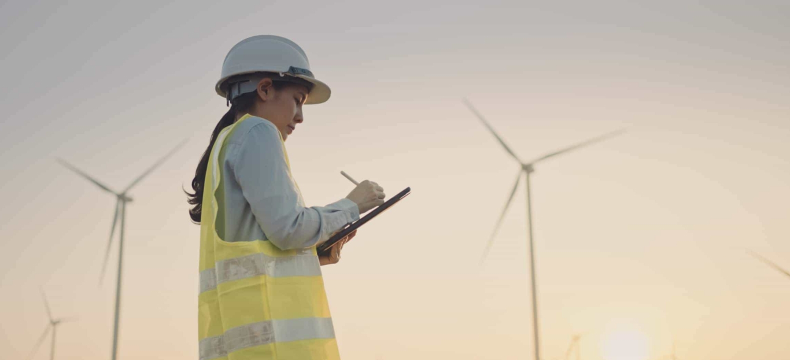 An engineer checks wind turbines and takes notes on their tablet.