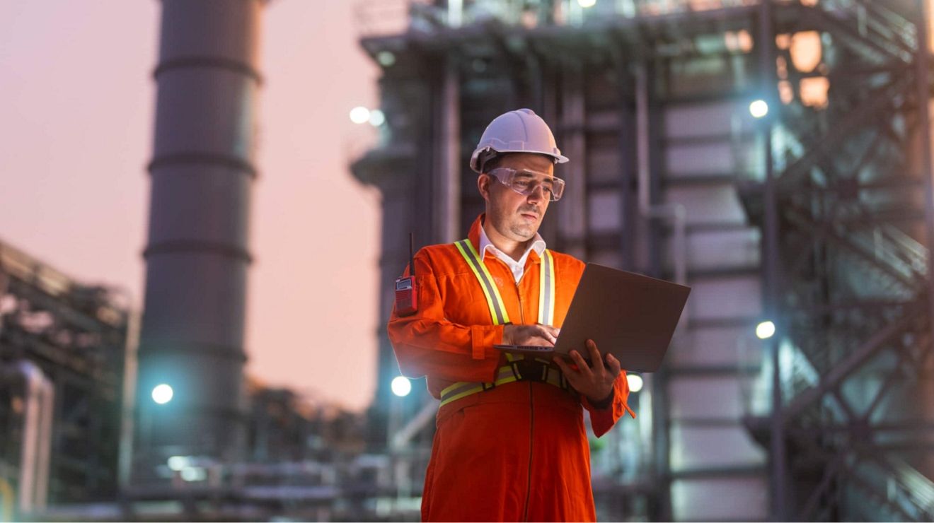 An engineer at an oil refinery studies their tablet while standing outside at dusk. 