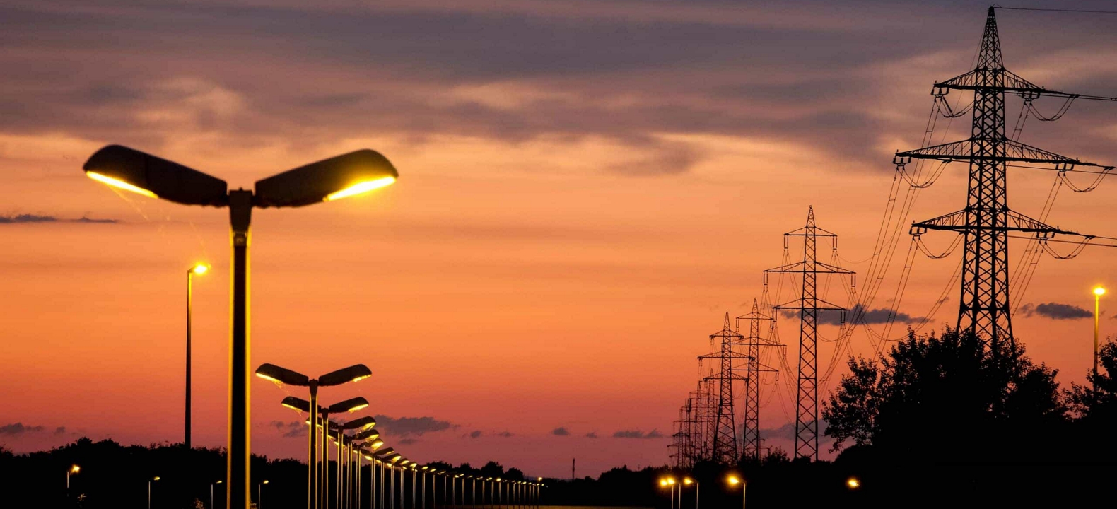 A row of high-voltage electrical towers alongside a row of streetlights shining at dusk.