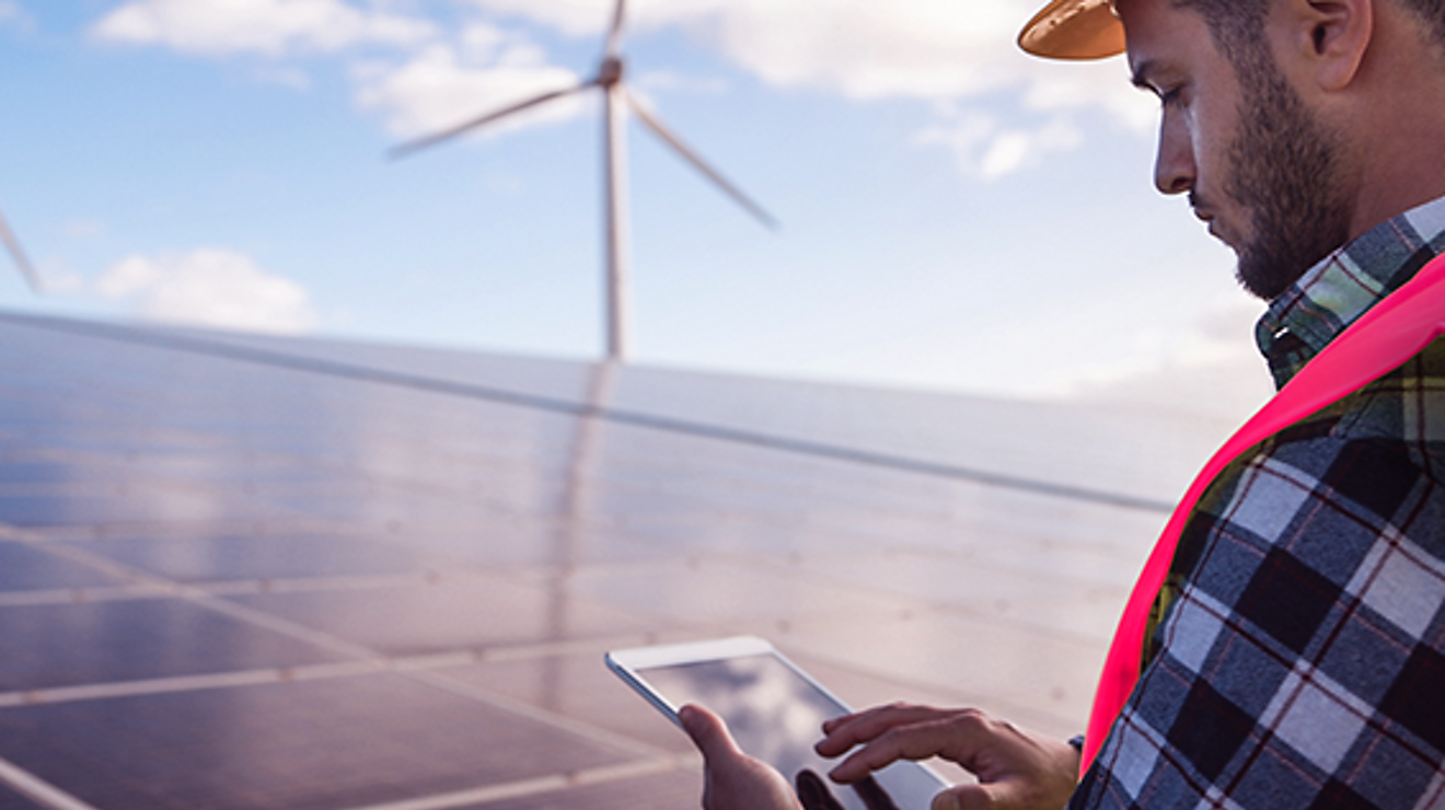 Worker with vest and hardhat looks at tablet in front of wind turbines