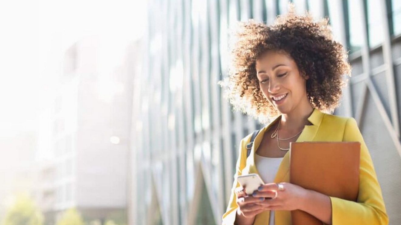 A smiling business professional works efficiently from their mobile device outside an office building.