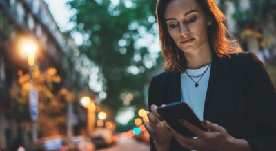 A financial services professional checks her smartphone while walking on a city street.