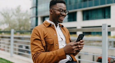 A smiling financial services professional uses his smartphone while walking across a business campus.