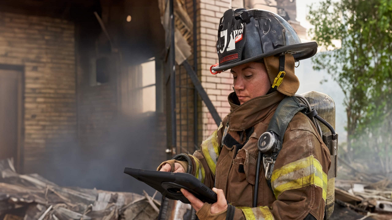A firefighter in protective gear works with a tablet within the wreckage of a burnt structure.
