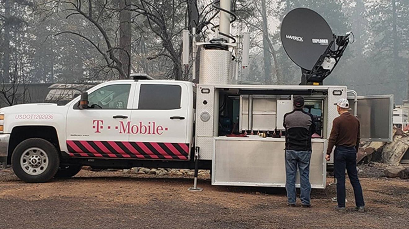 Two people inspect a T-Mobile emergency satellite internet truck at a natural disaster site near a wildfire.