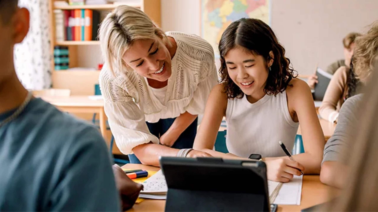 A woman teacher leaning over the shoulder of a female student using laptop.