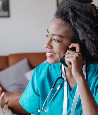 A doctor in scrubs and a stethoscope talks on her smartphone in a common area. 