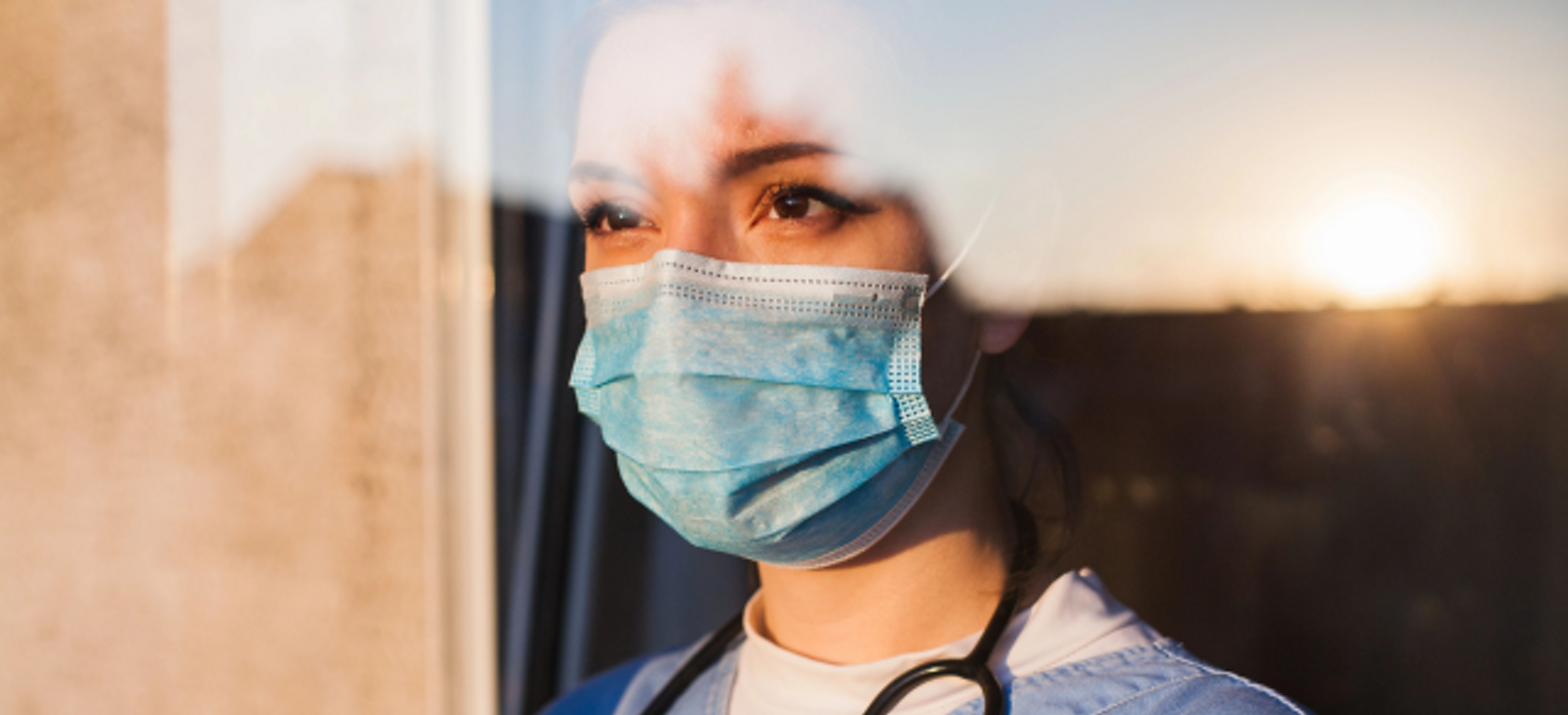 A masked medical professional gazes into the distance through a window.