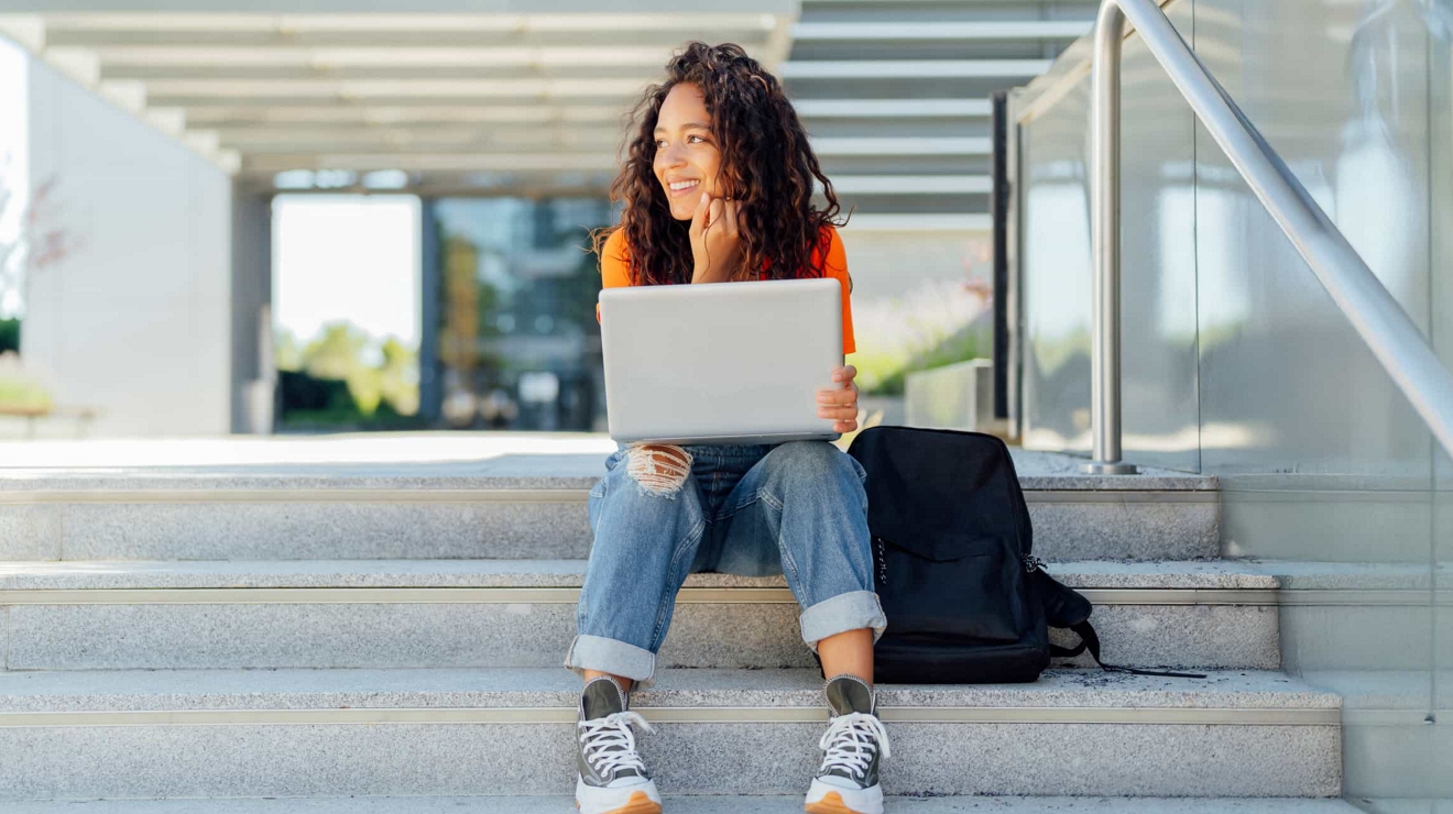 A happy college student sits outside a campus building with an open laptop resting on their knees.