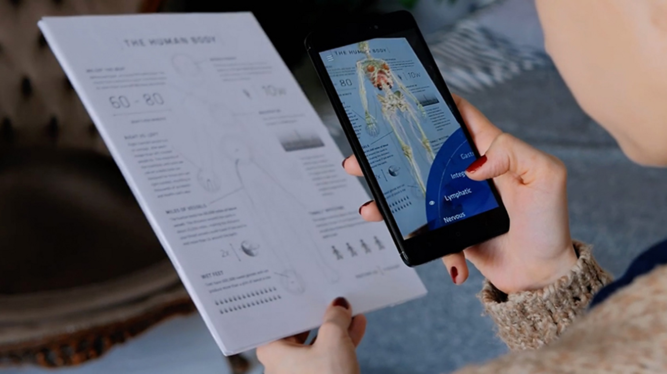 A hand holds a smartphone over a paper about the human body with a skeleton and anatomy appearing on screen