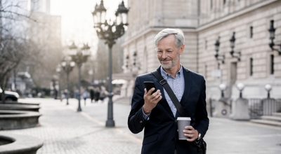 A professional traveling on business walks through a city with his smartphone and coffee in hand.