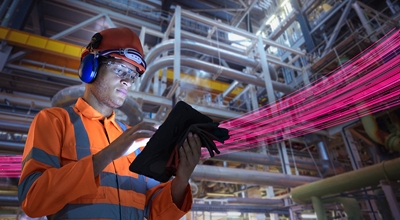 An industrial employee checks his tablet while magenta beams indicate connectivity across the plant.