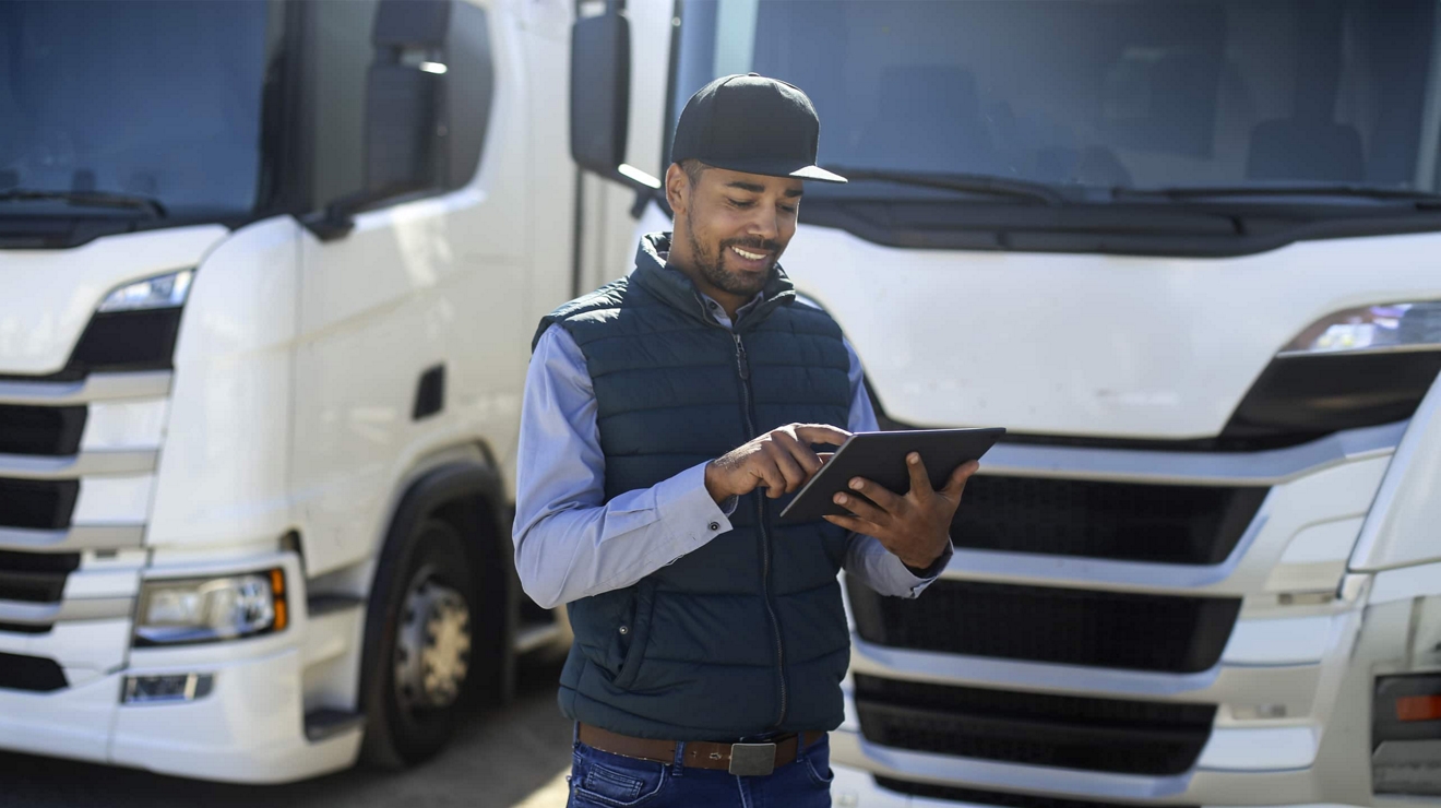 Smiling man uses a tablet while standing in front of two parked semi-trucks.